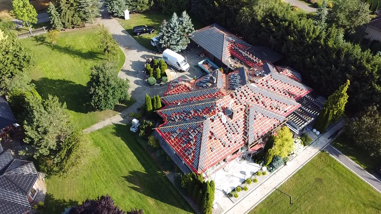 Widok z lotu ptaka na wielospadowy dach domu w trakcie wymiany pokrycia, widoczne fragmenty starego pokrycia, nowa więźba, samochód dostawczy i kontener na odpady na posesji.
