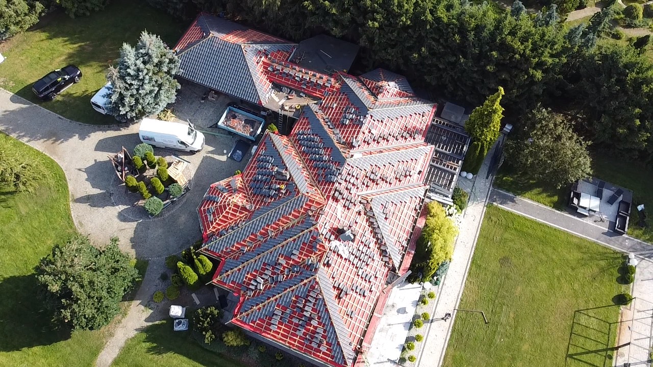Widok z lotu ptaka na rozległy dach w trakcie układania szarej dachówki na czerwonej membranie, z widocznymi elementami więźby dachowej, na tle zadbanego ogrodu z pojazdami i meblami ogrodowymi.