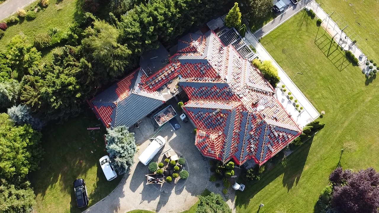 Widok z lotu ptaka na dom w trakcie wymiany pokrycia dachowego, widoczne elementy więźby i częściowo ułożone nowe dachówki, teren wokół domu zadbany z elementami architektury krajobrazu.