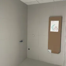 Wykończona łazienka z szarymi, kwadratowymi płytkami na ścianach, wystającymi rurami instalacyjnymi i kartonem z grafiką grzejnika Atlantic 2012 opartym o ścianę.