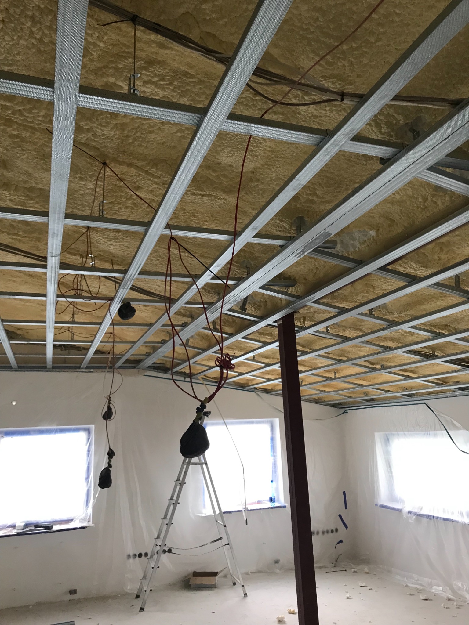 Sufit w trakcie izolacji z widoczną metalową konstrukcją nośną, przewodami elektrycznymi i oknami zabezpieczonymi folią malarską. W pomieszczeniu stoi drabina. Widoczne ślady prac remontowych.
