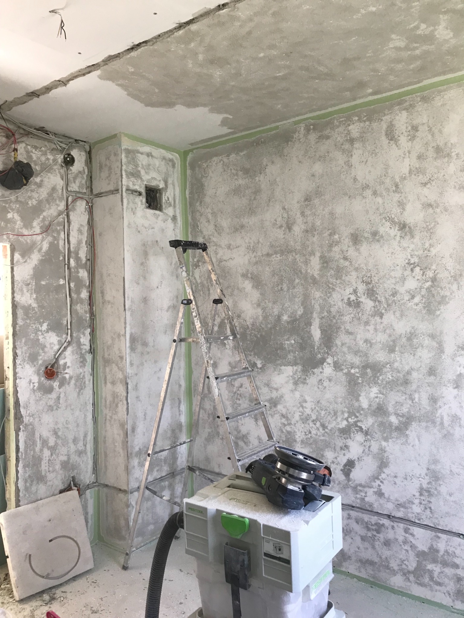 Ściana w trakcie przygotowywania pod wykończenie, z widoczną strukturą, zabezpieczona taśmą malarską, drabina aluminiowa i szlifierka do gładzi na odkurzaczu przemysłowym.