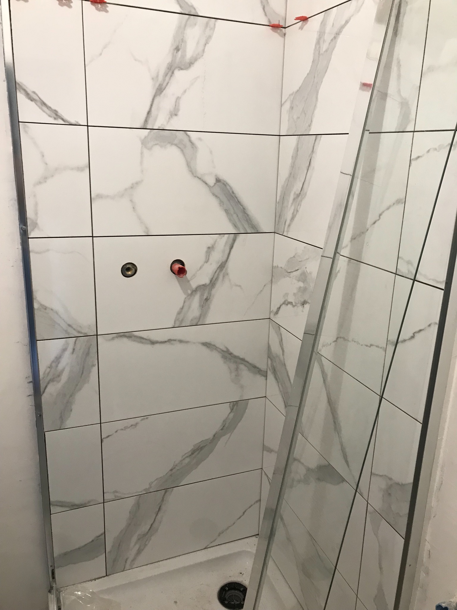 Ściana kabiny prysznicowej w trakcie wykańczania, widoczne białe płytki imitujące marmur z szarymi żyłami, elementy montażowe i czerwone kliny dystansowe.