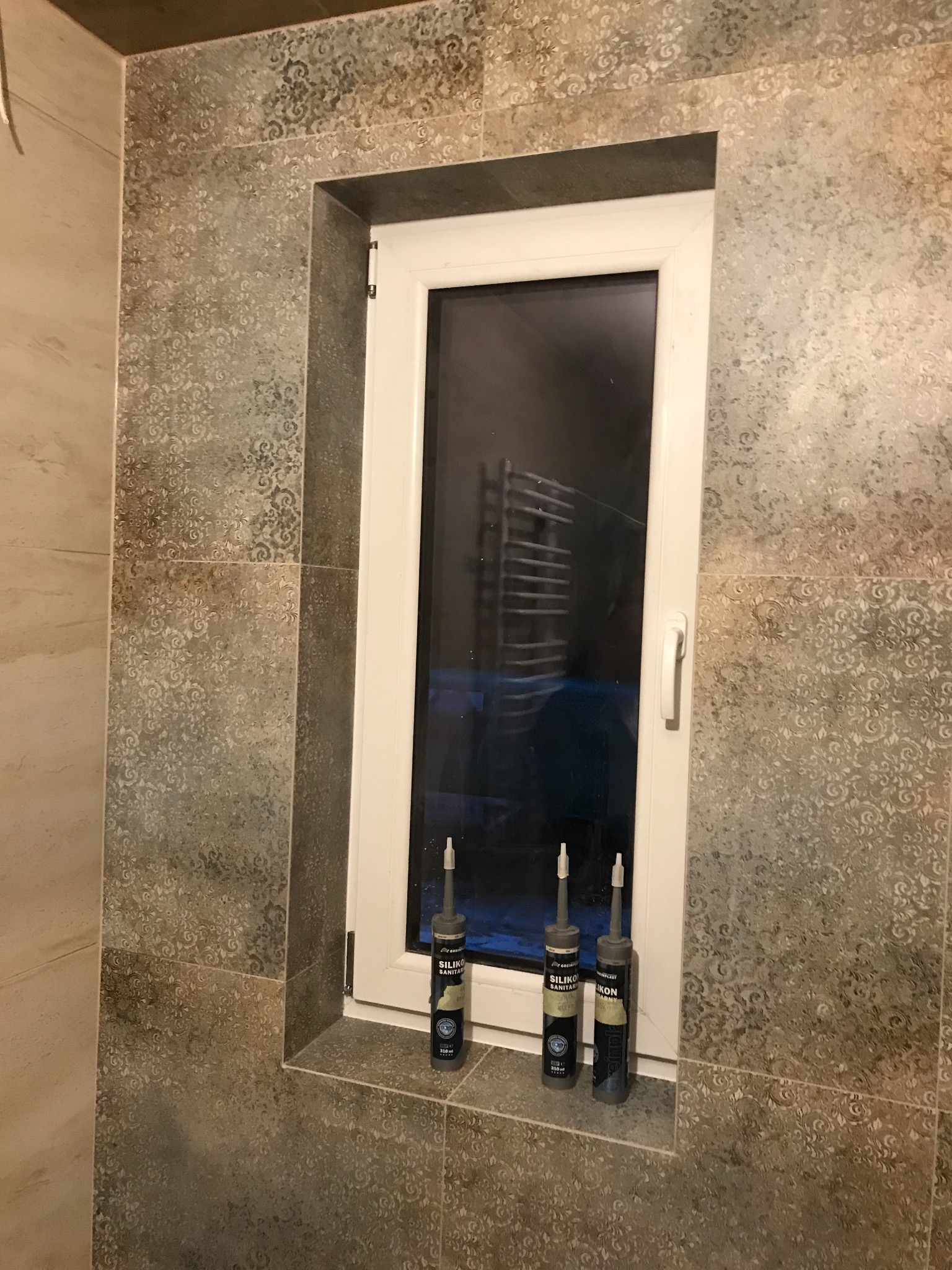 Błyszczące, wzorzyste płytki ceramiczne w łazience, obramowujące okno z widocznym grzejnikiem, z trzema tubami silikonu sanitarnego postawionymi na parapecie okiennym.