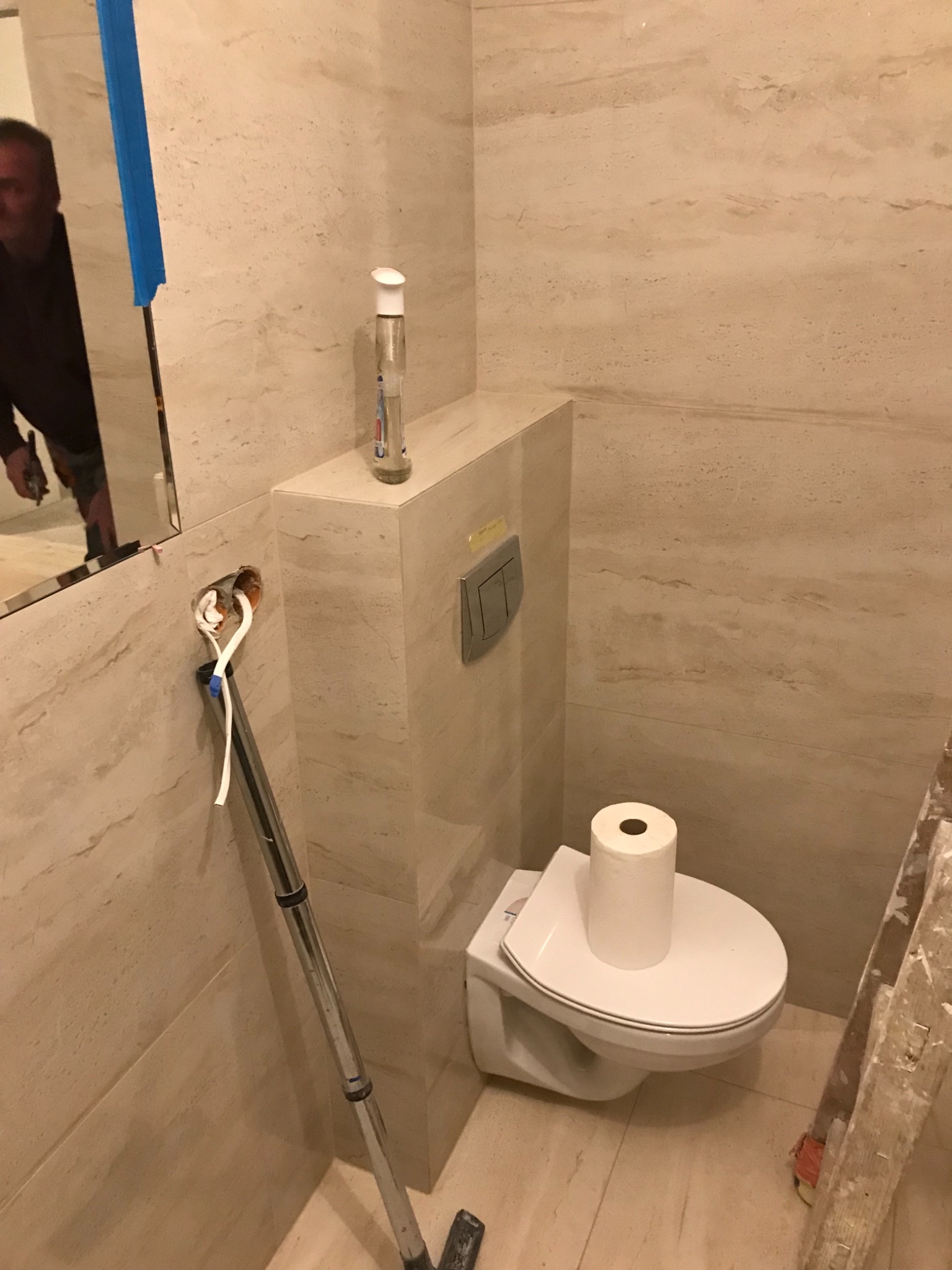 Wnętrze łazienki w trakcie wykańczania: beżowe płytki imitujące marmur, wisząca toaleta z rolką papieru na desce, widoczne przewody elektryczne przy lustrze, drabina malarska oparta o ścianę.
