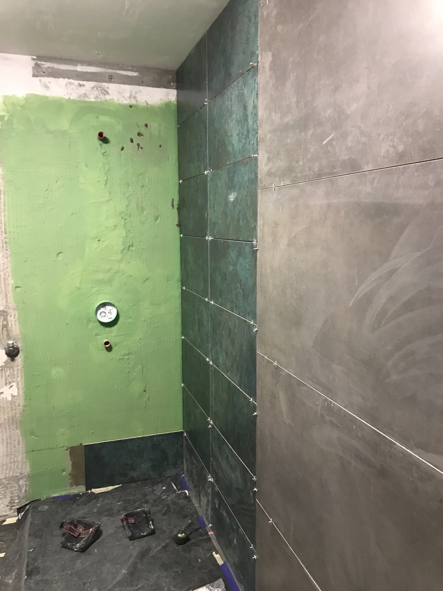 Fragment ściany w trakcie kładzenia płytek w łazience, widoczne krzyżyki dystansowe, zielona ściana z wystającymi rurami, narzędzia na podłodze.