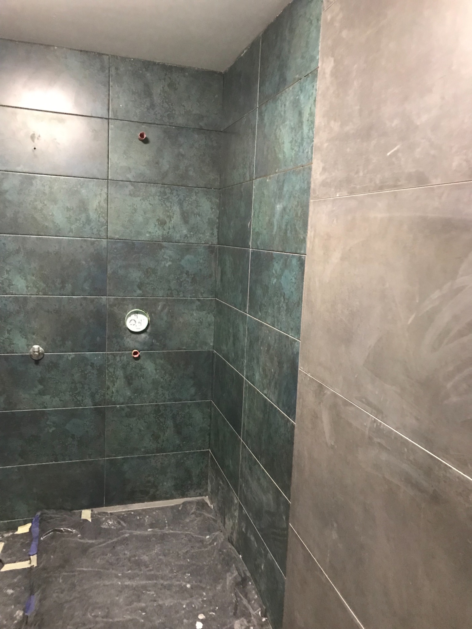 Fragment łazienki w trakcie prac wykończeniowych; widoczne płytki w odcieniach zieleni i szarości, zabezpieczona folią podłoga oraz wystające elementy instalacji.