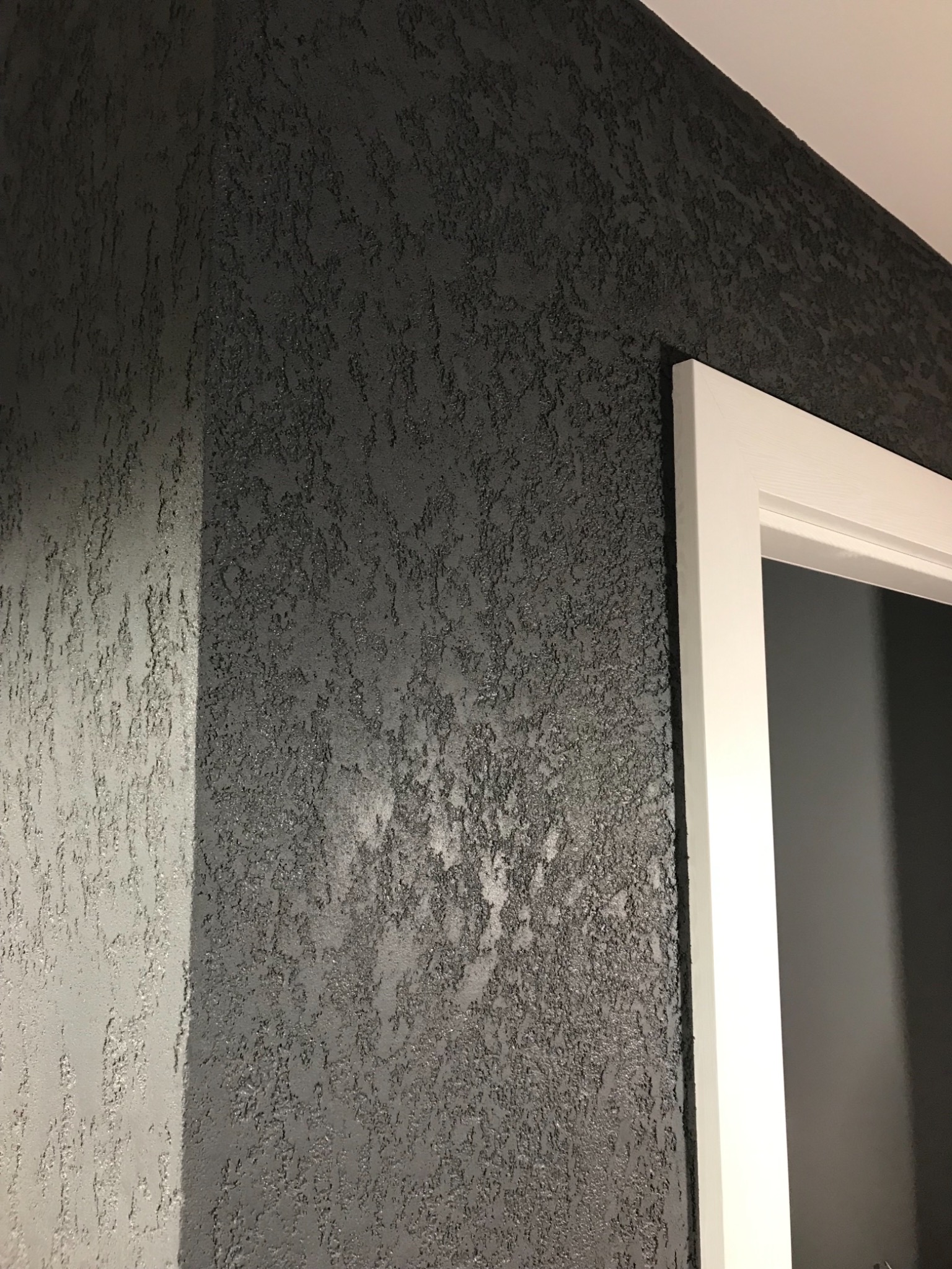 Ciemnoszara ściana z wyraźną, ziarnistą fakturą, oświetlona z boku, sąsiadująca z białą ościeżnicą drzwiową.