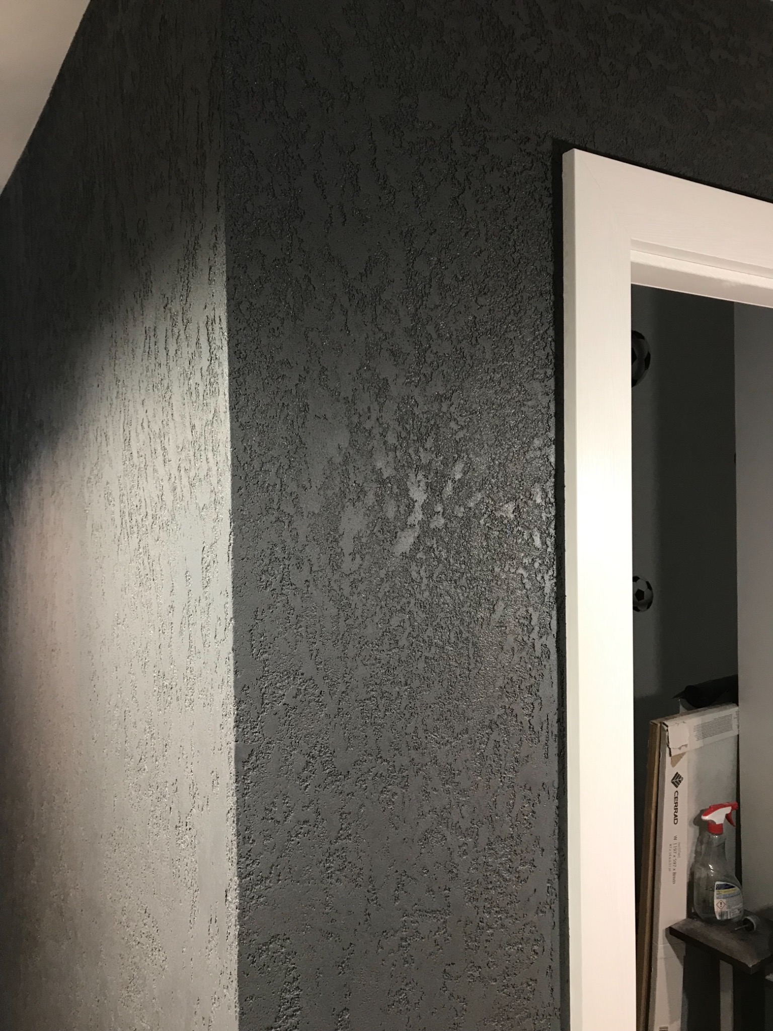 Szara ściana z wyraźną fakturą, oświetlona z lewej strony, przylegająca do białej futryny drzwi, widoczny fragment wnętrza z piłkami do piłki nożnej na ścianie i kartonem płytek podłogowych.