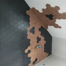 Ściana łazienki wyłożona heksagonalnymi płytkami w dwóch odcieniach: grafitowym i drewnopodobnym, z widocznym fragmentem instalacji wodnej i białymi płytkami podłogowymi.