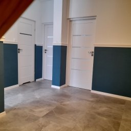 Łukasz - Wnętrze korytarza z trzema białymi drzwiami z poziomymi przeszkleniami, dolna część ścian pomalowana na ciemny granat, szare płytki podłogowe imitujące marmur.