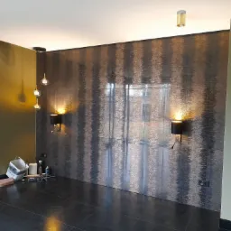 Ściana wykończona dekoracyjnymi płytkami z oświetleniem ściennym, widoczne narzędzia i akcesoria glazurnicze na podłodze z ciemnych płytek.