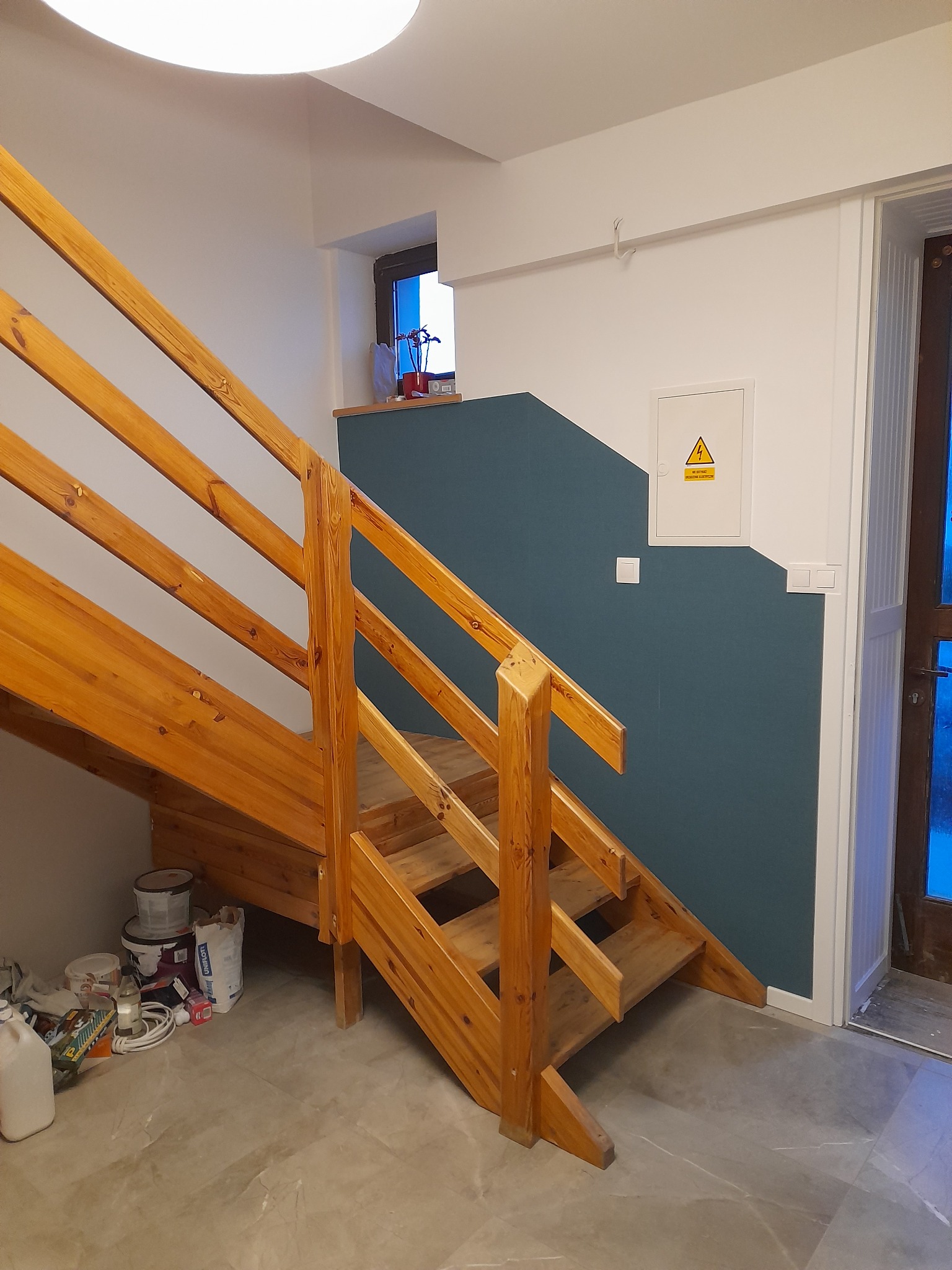 Drewniane schody w trakcie montażu, widoczne puszki z farbą i materiały budowlane na podłodze, ściana pomalowana na dwa kolory, biały i ciemny niebieski.