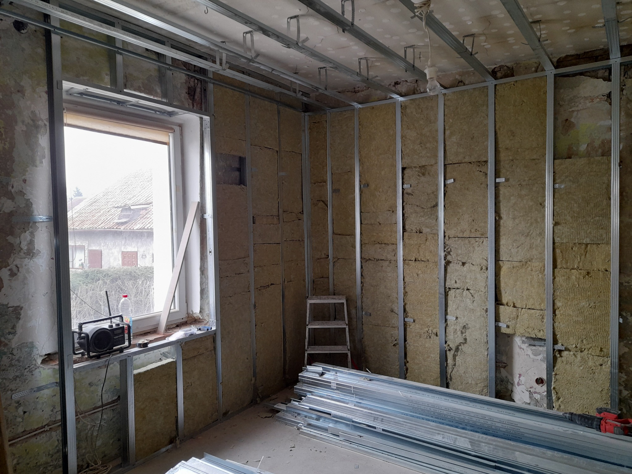 Wnętrze w trakcie prac remontowych: izolacja ścian wełną mineralną zamocowaną w metalowym stelażu, widoczny fragment starej ściany, radio na parapecie i stos profili metalowych na podłodze.