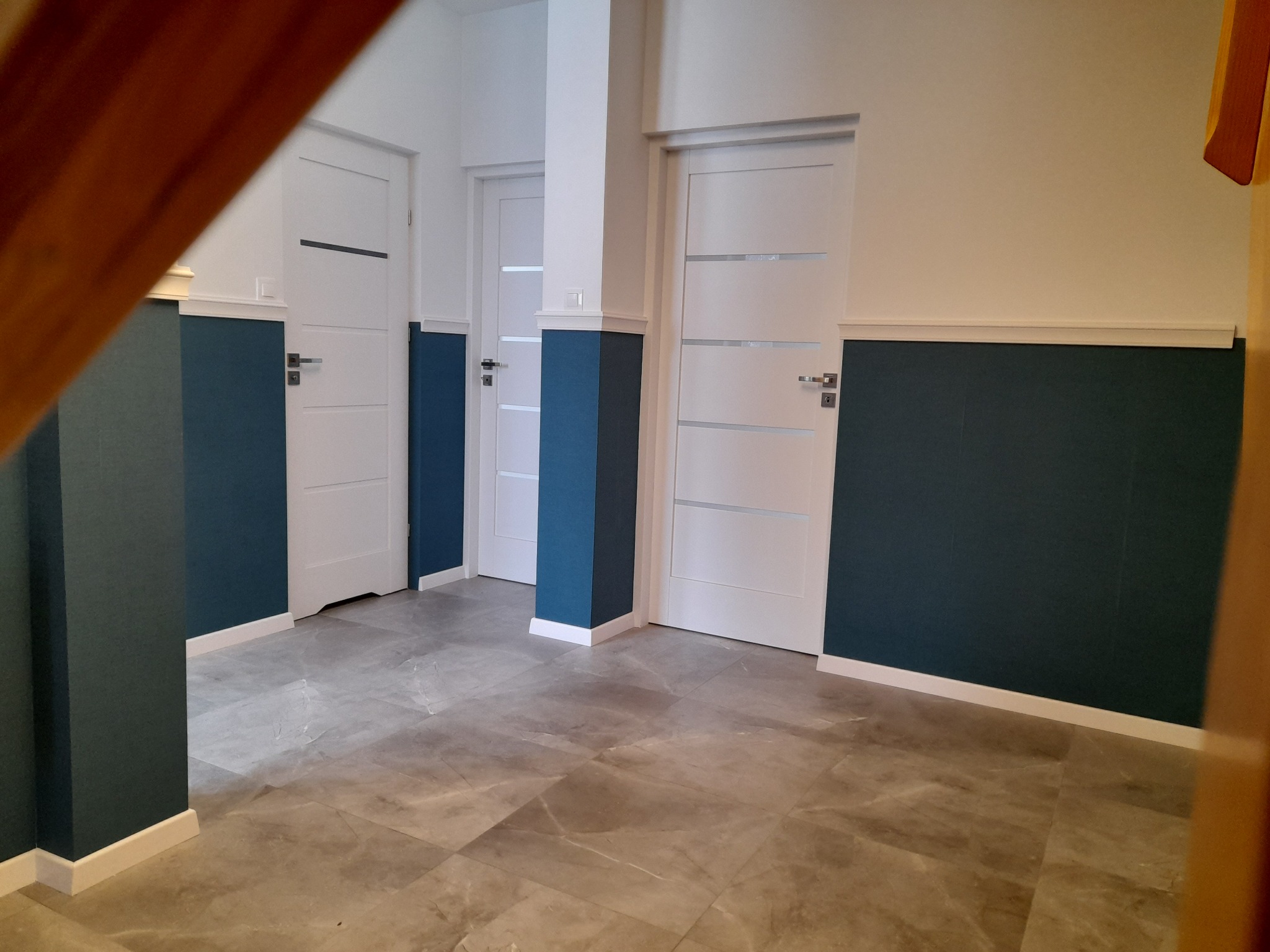 Wnętrze korytarza z trzema białymi drzwiami z poziomymi przeszkleniami, dolna część ścian pomalowana na ciemny granat, szare płytki podłogowe imitujące marmur.