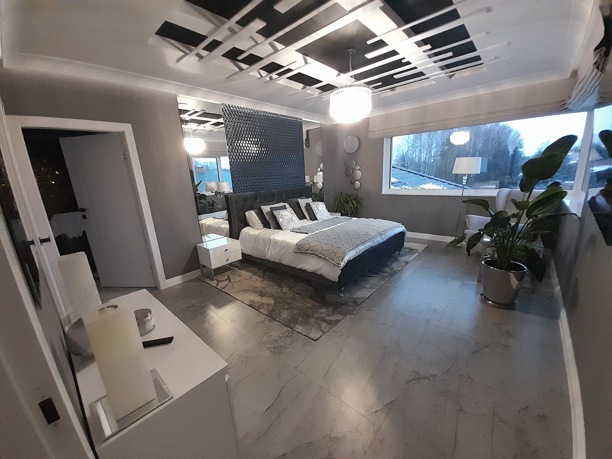 Sypialnia w Szczytnie z łóżkiem z pikowanym zagłówkiem, geometrycznym wzorem na suficie i marmurową podłogą, oświetlona wiszącą lampą i światłem z okna.