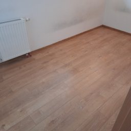 Michał Charewicz - Usługi Remontowe - Pokój po remoncie z panelami podłogowymi w odcieniach brązu, biały grzejnik na ścianie i białe ściany z widocznymi śladami po pracach malarskich.