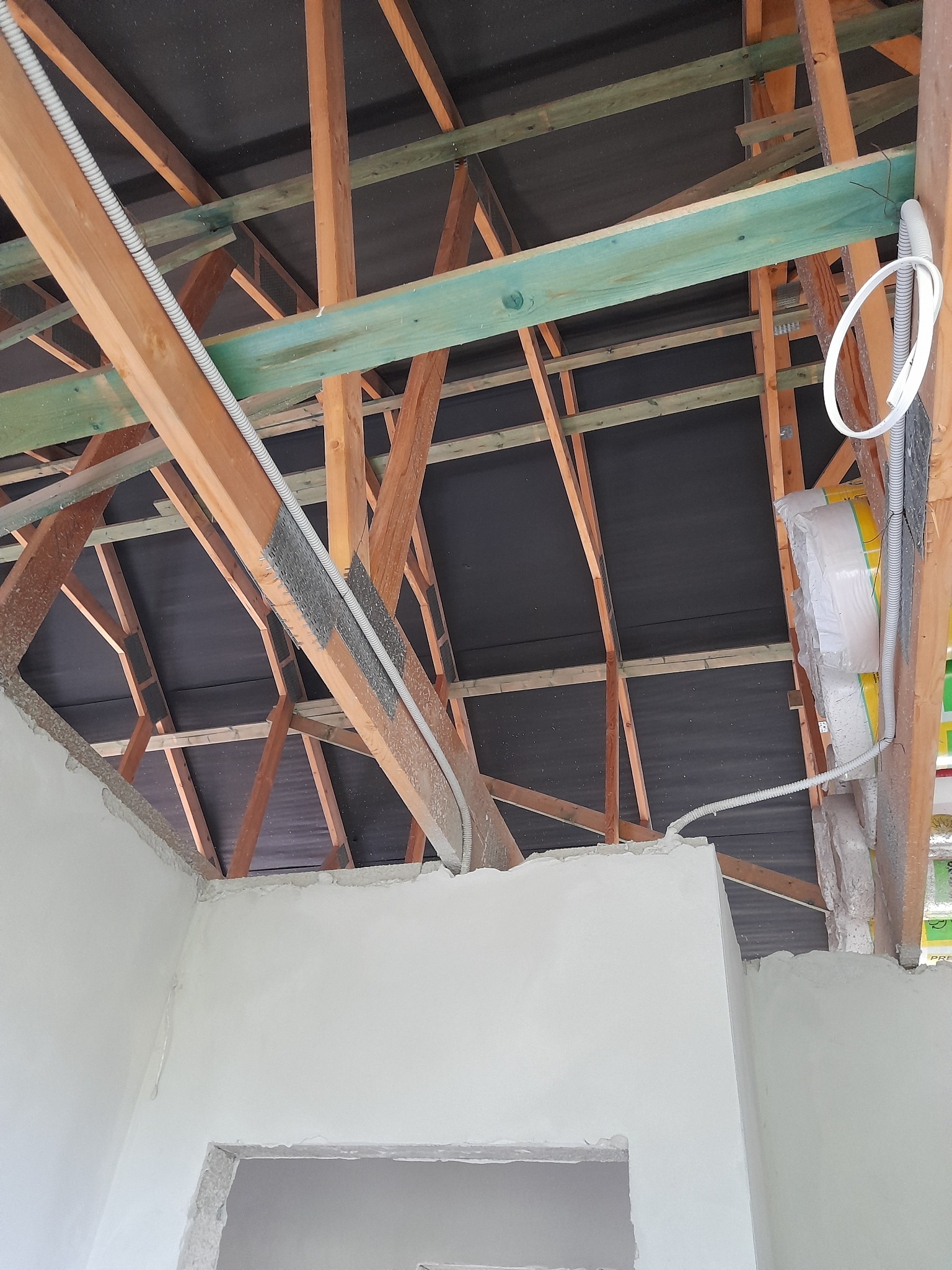 Surowy stan remontu poddasza: drewniana konstrukcja dachu z widoczną membraną, instalacja elektryczna w rurkach, ocieplenie w folii i częściowo otynkowane ściany z surowymi otworami drzwiowymi.