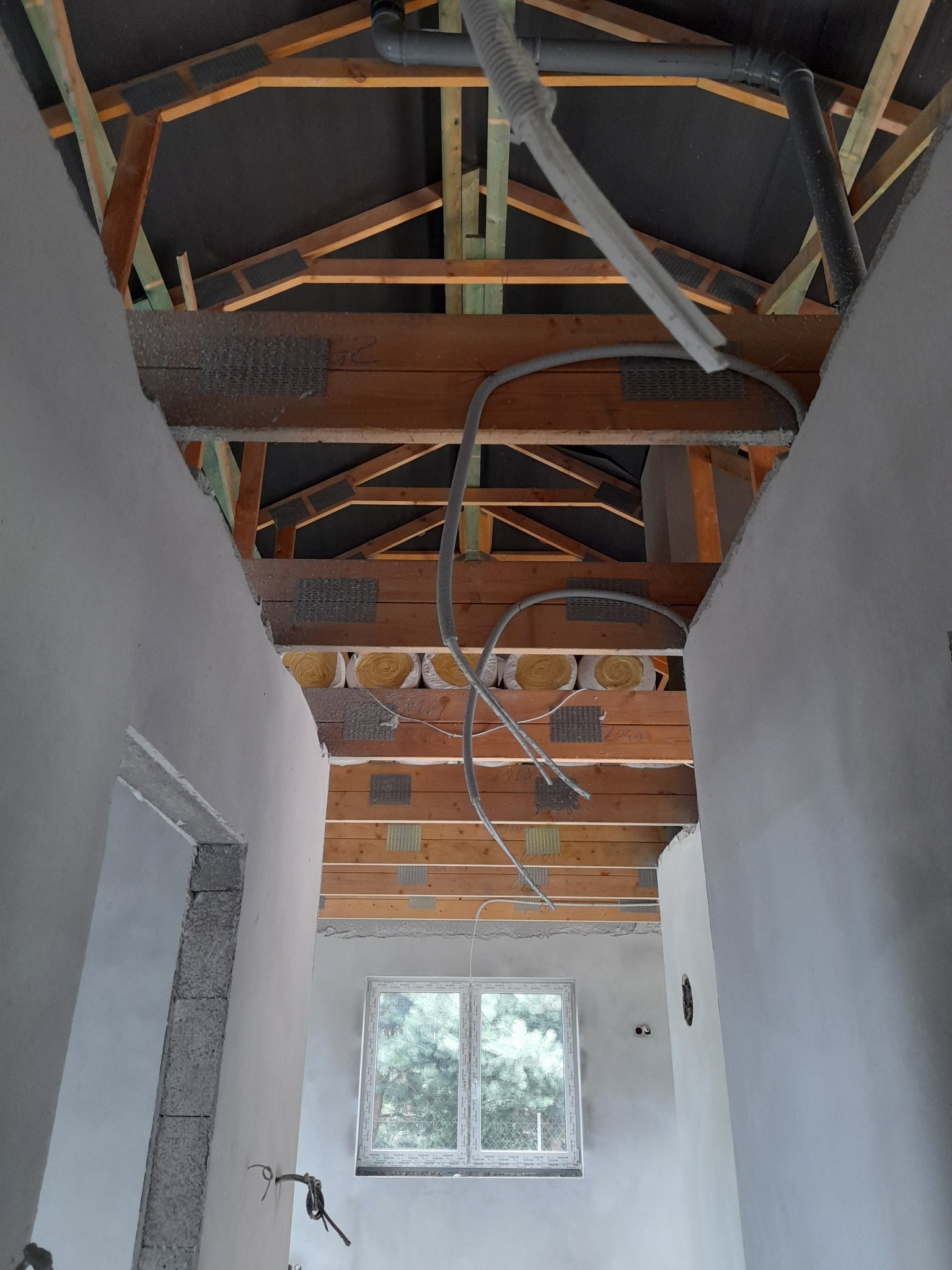 Wnętrze w trakcie prac wykończeniowych: surowa ściana, widoczna konstrukcja dachu z izolacją, okno z widokiem na drzewa, instalacje elektryczne i rury pod sufitem.