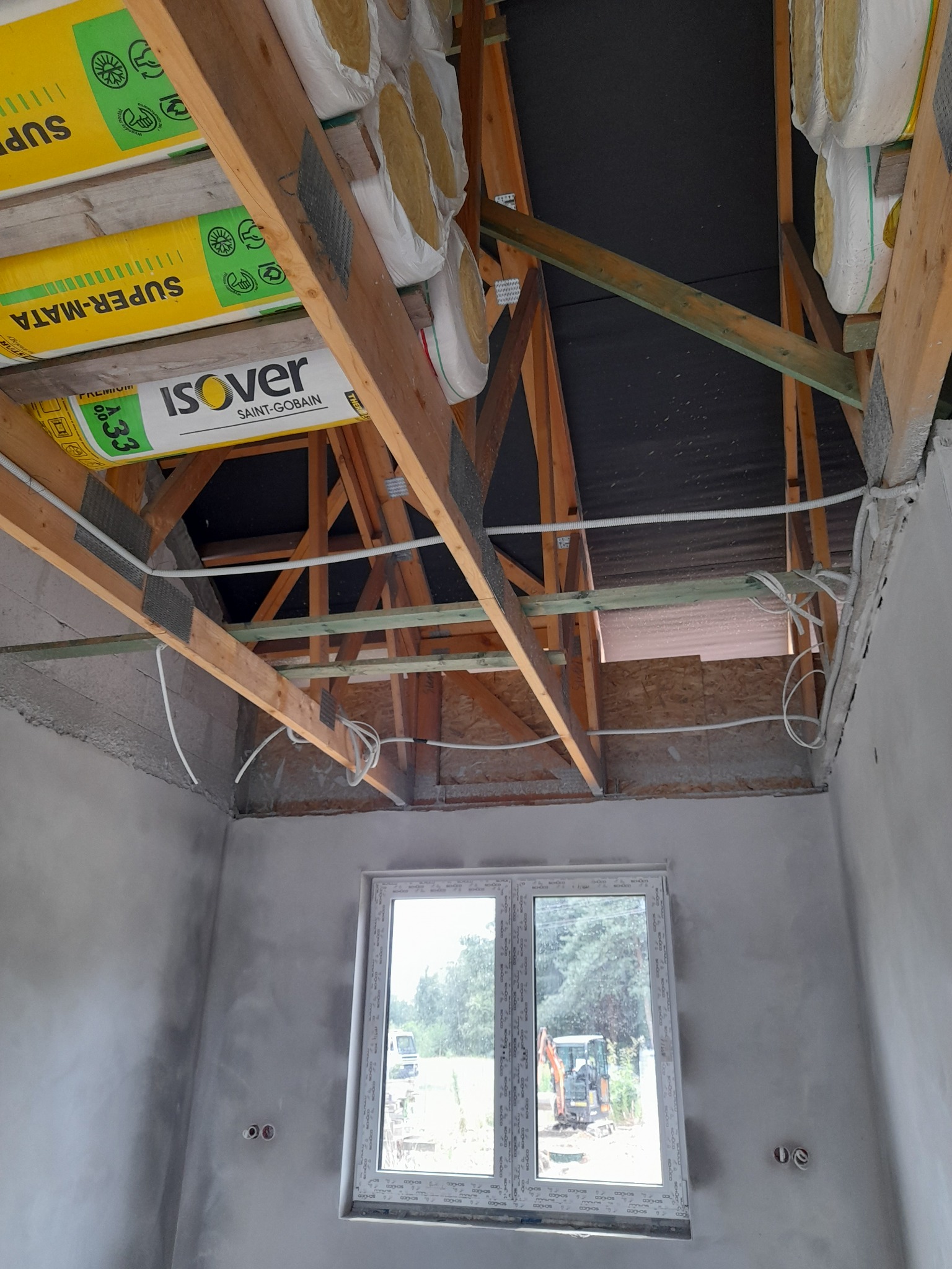 Poddasze w trakcie ocieplania, widoczne drewniane krokwie, izolacja z wełny mineralnej Isover Super-Mata A33, instalacja elektryczna w peszlach i surowe, otynkowane ściany z oknem.