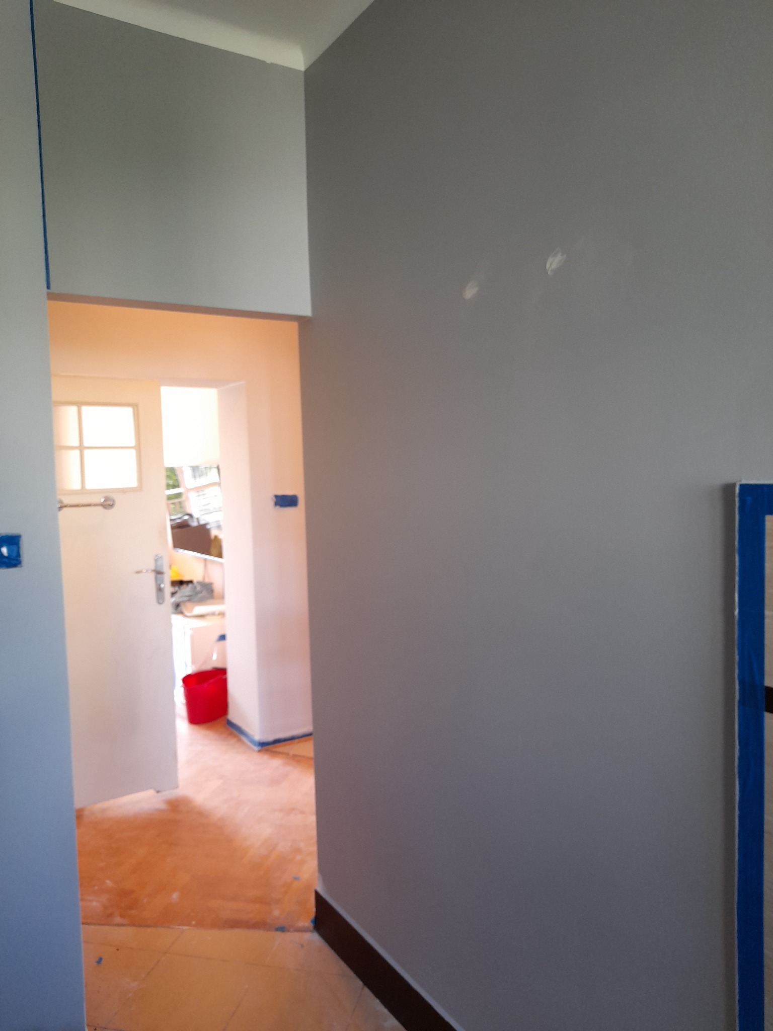 Świeżo pomalowana szara ściana z niebieską taśmą malarską przy suficie i podłodze, widok na otwarte drzwi do pokoju z czerwoną miską i pomalowanymi na beżowo ścianami.