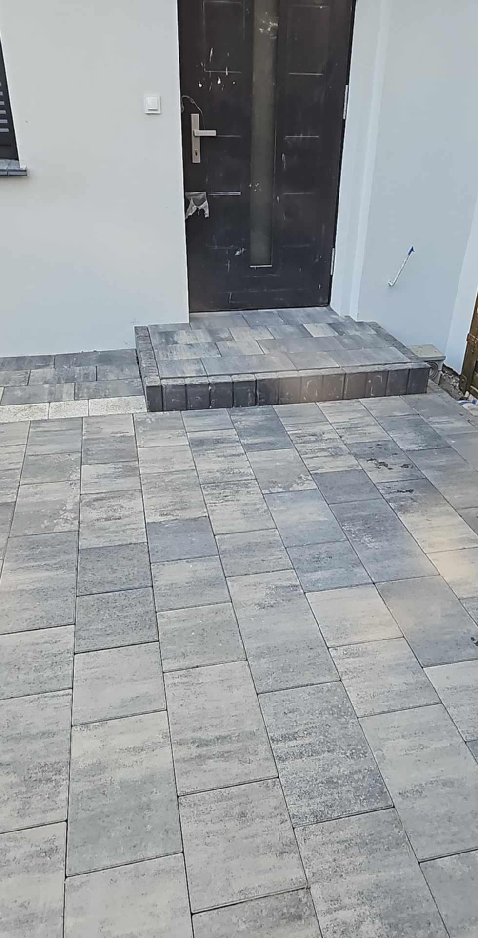 Ułożona szara kostka brukowa przed wejściem do domu z czarnymi drzwiami i schodami z kostki.