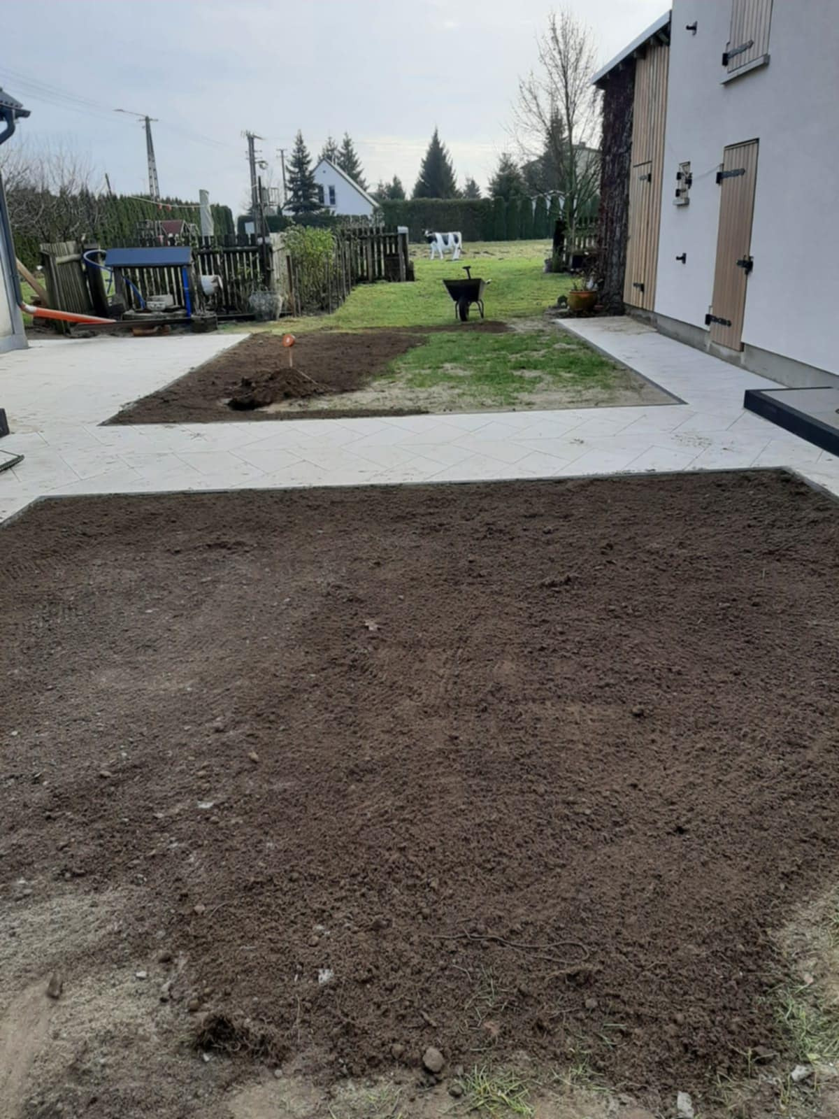 Przygotowany teren pod budowę ścieżki z kostki, widoczne wytyczone obszary i obrzeża z jasnej kostki. W tle trawnik, taczka i dekoracyjna figura krowy.