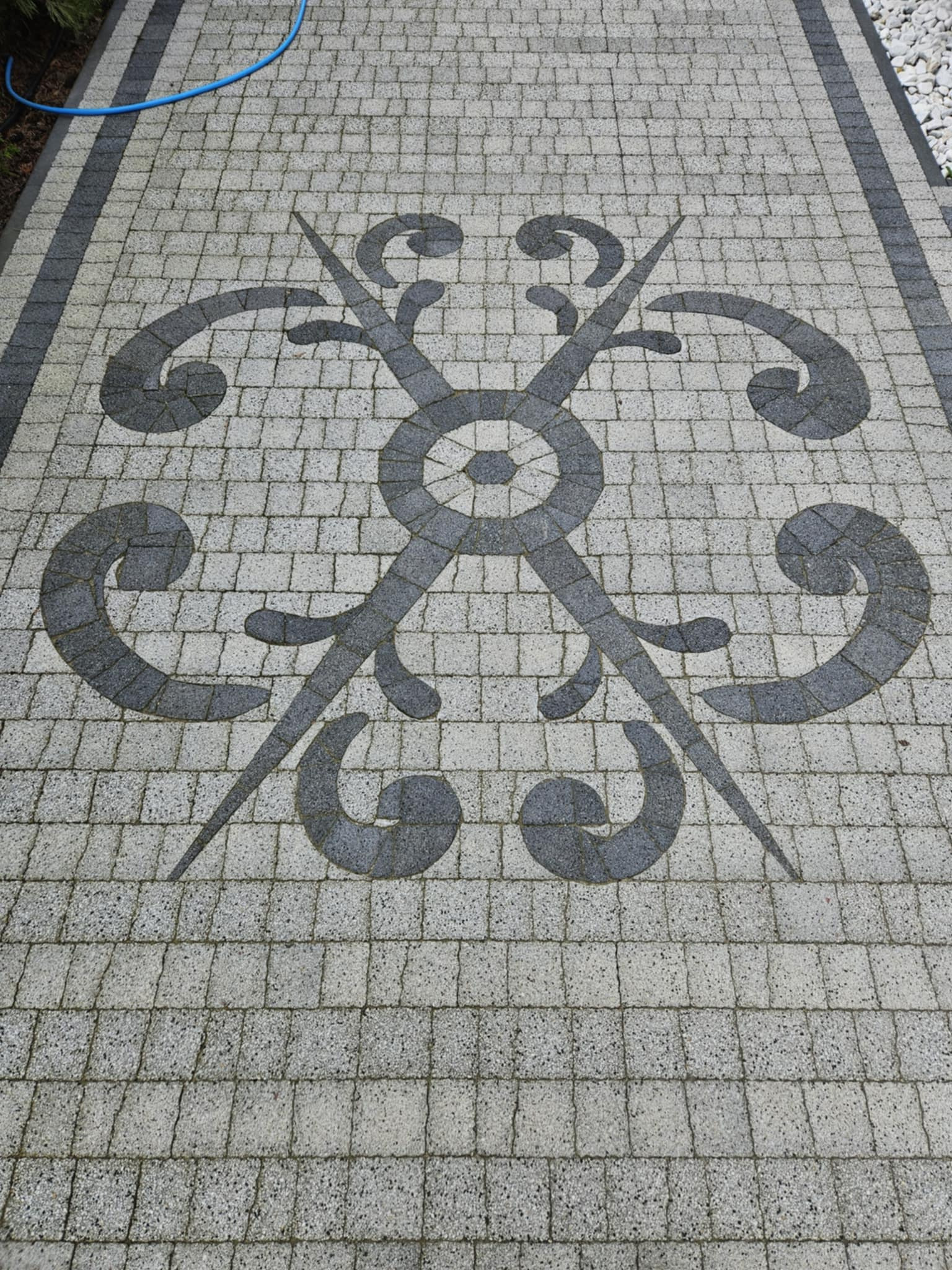 Wzór dekoracyjny ułożony z kostki brukowej w odcieniach szarości, otoczony jasną kostką i obrzeżem. W tle widoczny wąż ogrodowy i białe kamienie.