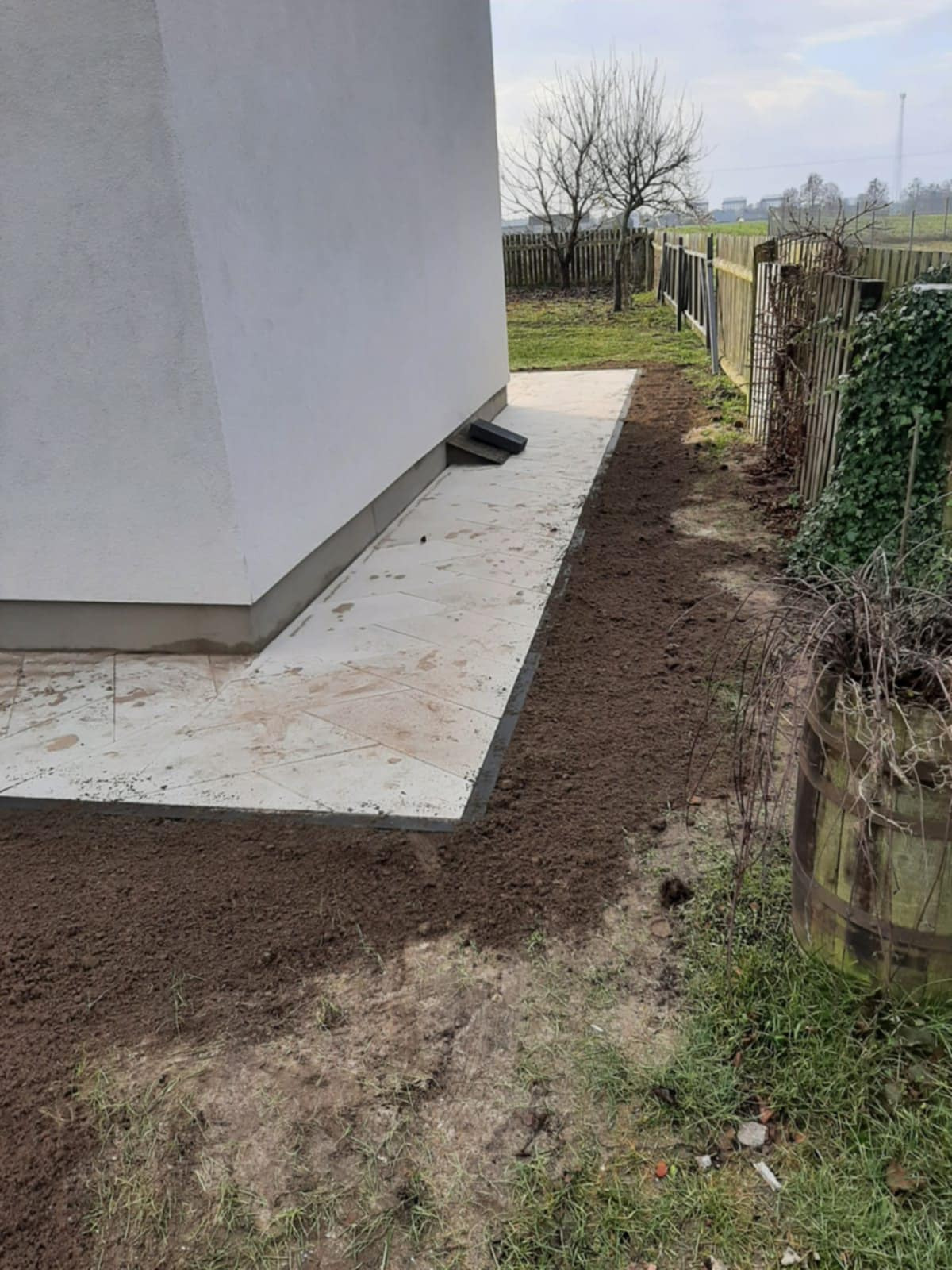 Wyłożony jasnymi płytami chodnikowymi teren wokół budynku, obsypany świeżą ziemią, widok z boku, w tle drewniany płot i drzewa bez liści.