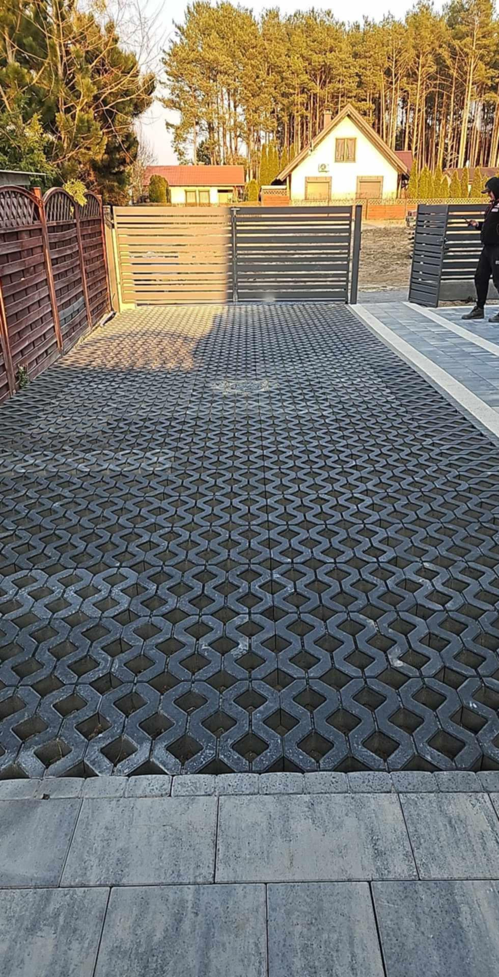 Szara, ażurowa kostka brukowa ułożona na podjeździe przed posesją z brązowym i szarym ogrodzeniem oraz dwuskrzydłową bramą w tle, widoczny fragment domu i drzewa.