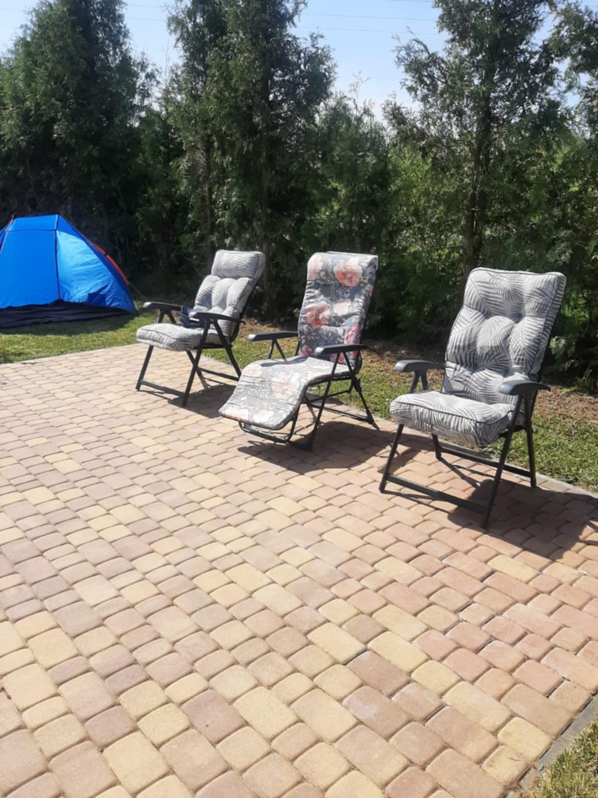 Ułożona kostka brukowa w odcieniach beżu i brązu, z trzema rozłożonymi fotelami ogrodowymi z poduszkami w tle i niebieskim namiotem w oddali.