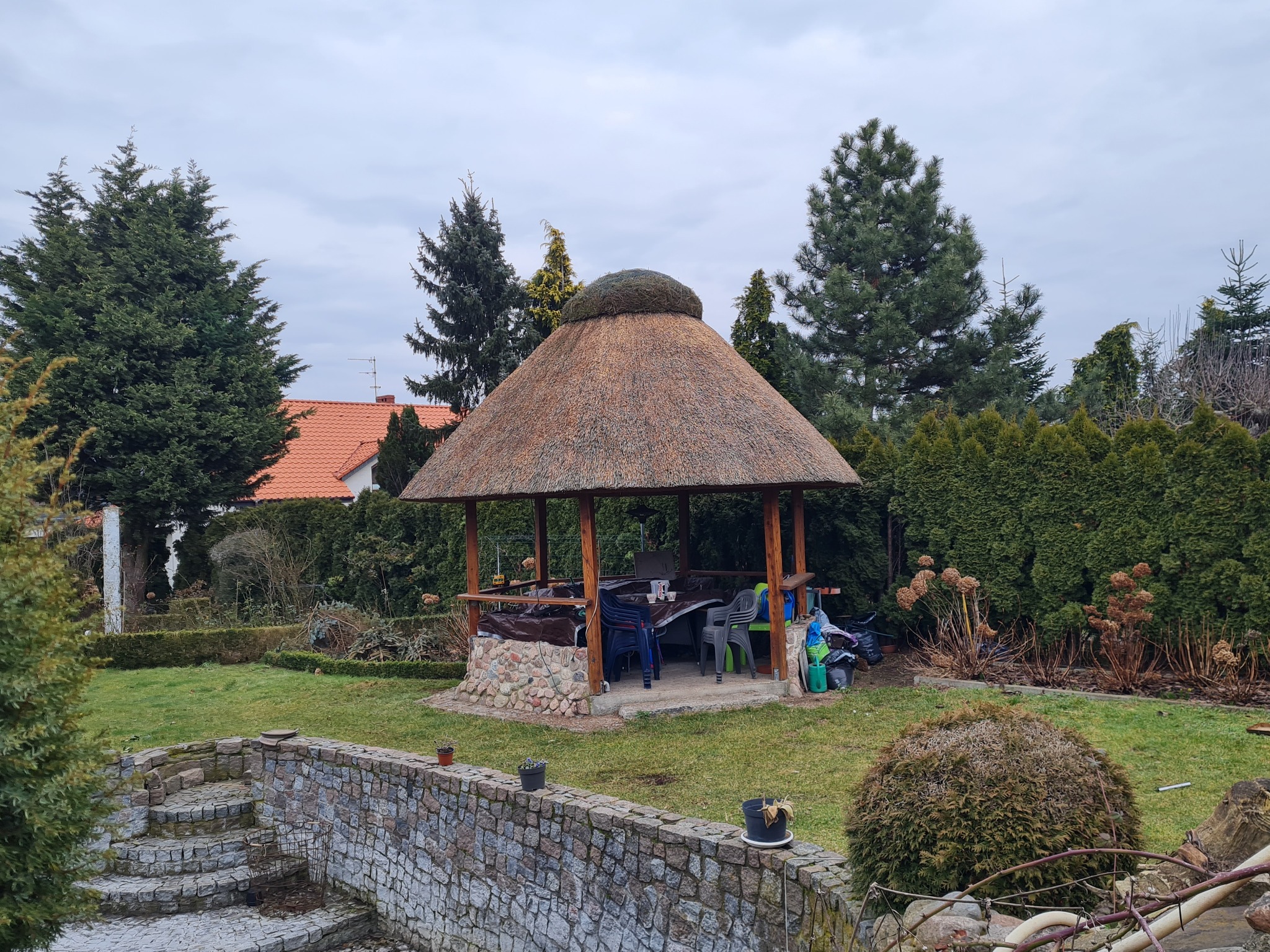 Altana z dachem krytym strzechą, otoczona zielenią ogrodu, z widocznymi schodami z kamienia i elementami architektury ogrodowej w tle.