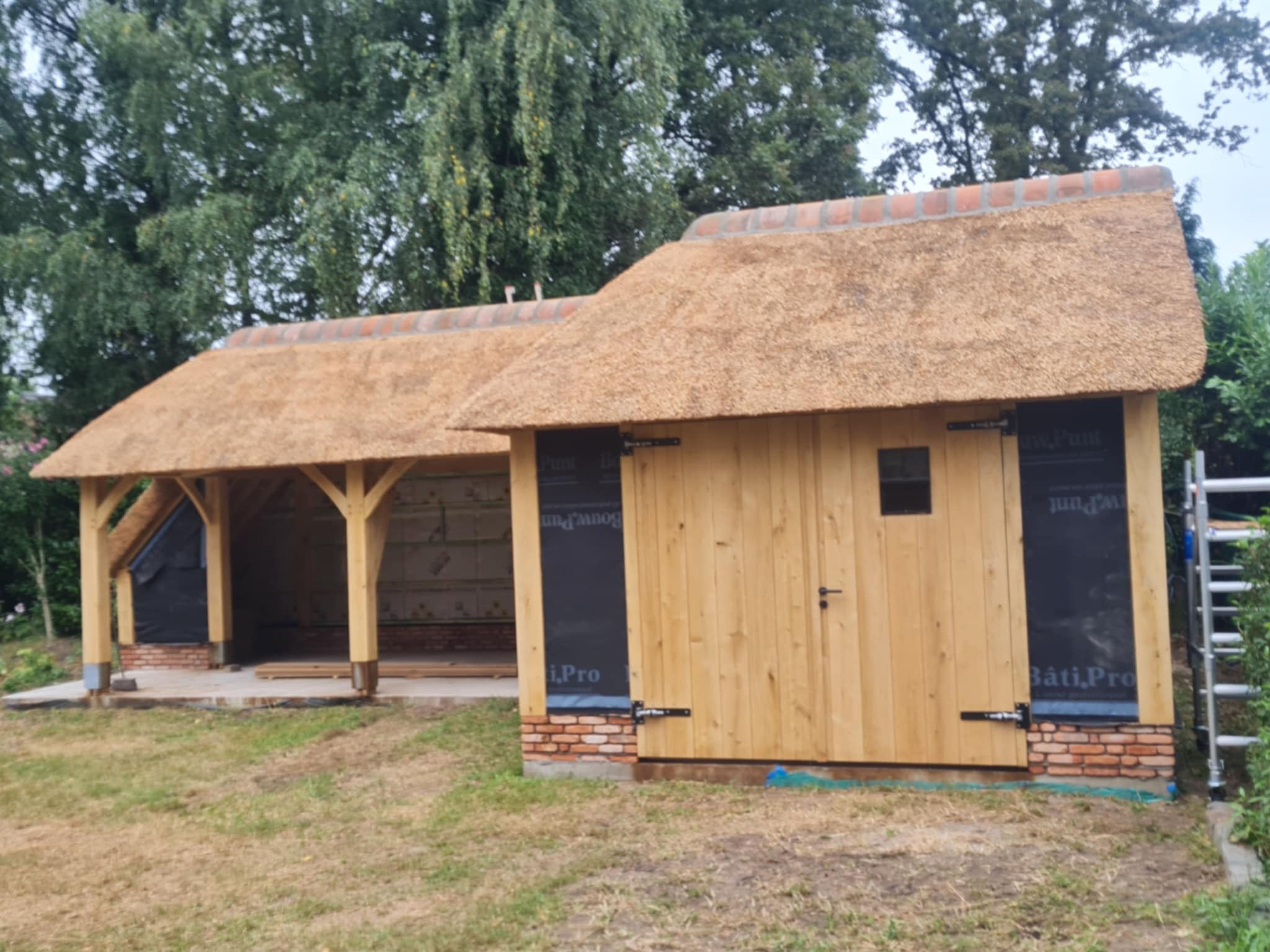 Nowo wybudowana drewniana szopa z dachem krytym strzechą i ceglaną podmurówką, widoczne materiały izolacyjne i rusztowanie w tle.