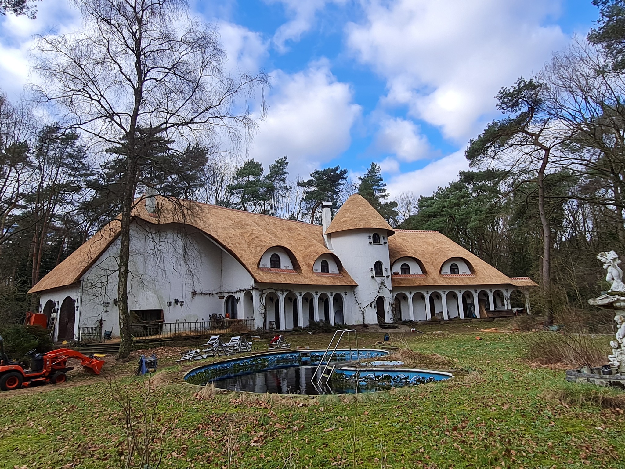 Rozległy dom z białymi ścianami i strzechą, widoczny z ogrodu z zaniedbanym basenem i małym traktorem, na tle lasu i pochmurnego nieba.