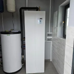 Biała pompa ciepła marki York, zbiornik akumulacyjny ciepłej wody użytkowej, instalacja hydrauliczna w szarym pomieszczeniu z szarą podłogą i okładziną ścienną imitującą drewno.