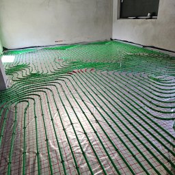 AQUA GREEN Usługi hydrauliczne Dawid Karolczyk - Rozłożona instalacja ogrzewania podłogowego z zielonych rurek na folii z czerwoną kratką w surowym pomieszczeniu przed wylewką.