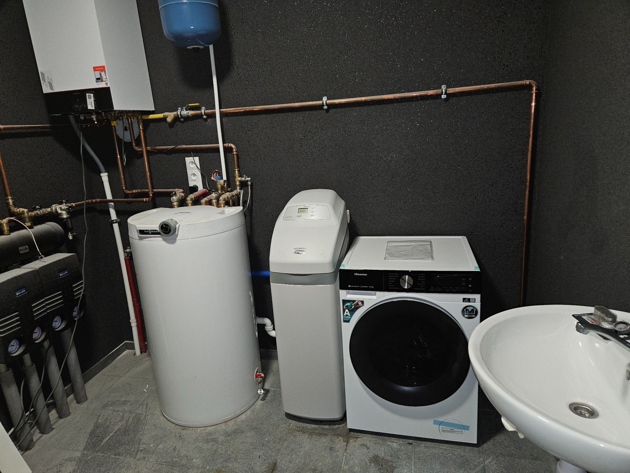 Kotłownia w piwnicy z piecem gazowym, zasobnikiem ciepłej wody, zmiękczaczem wody, pralką i umywalką na tle ściany z czarnego granulatu.