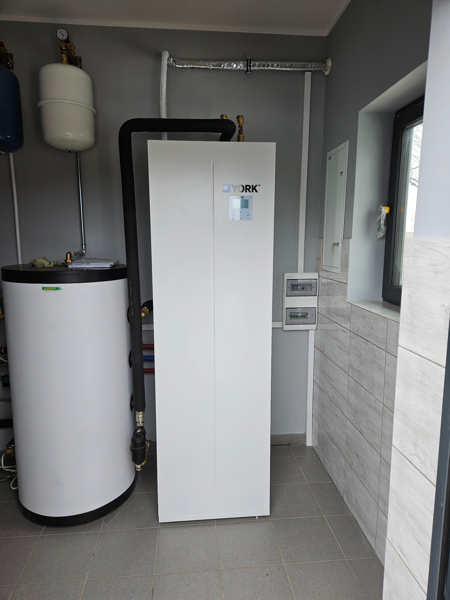 Biała pompa ciepła marki York, zbiornik akumulacyjny ciepłej wody użytkowej, instalacja hydrauliczna w szarym pomieszczeniu z szarą podłogą i okładziną ścienną imitującą drewno.
