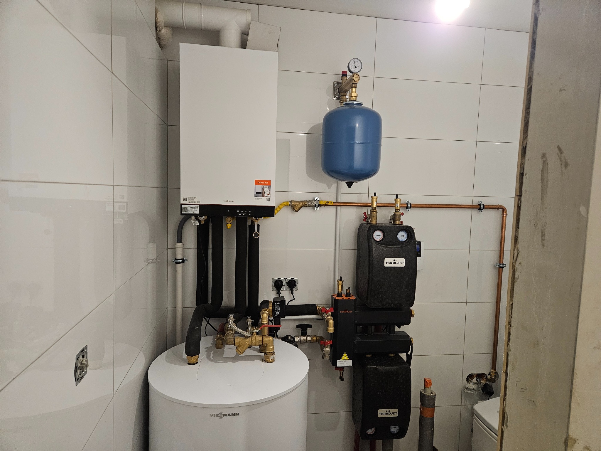Nowoczesna kotłownia z białym piecem gazowym Viessmann, niebieskim zbiornikiem wyrównawczym i czarnym zestawem pompowym Termojet, zamontowana na białych, błyszczących płytkach.