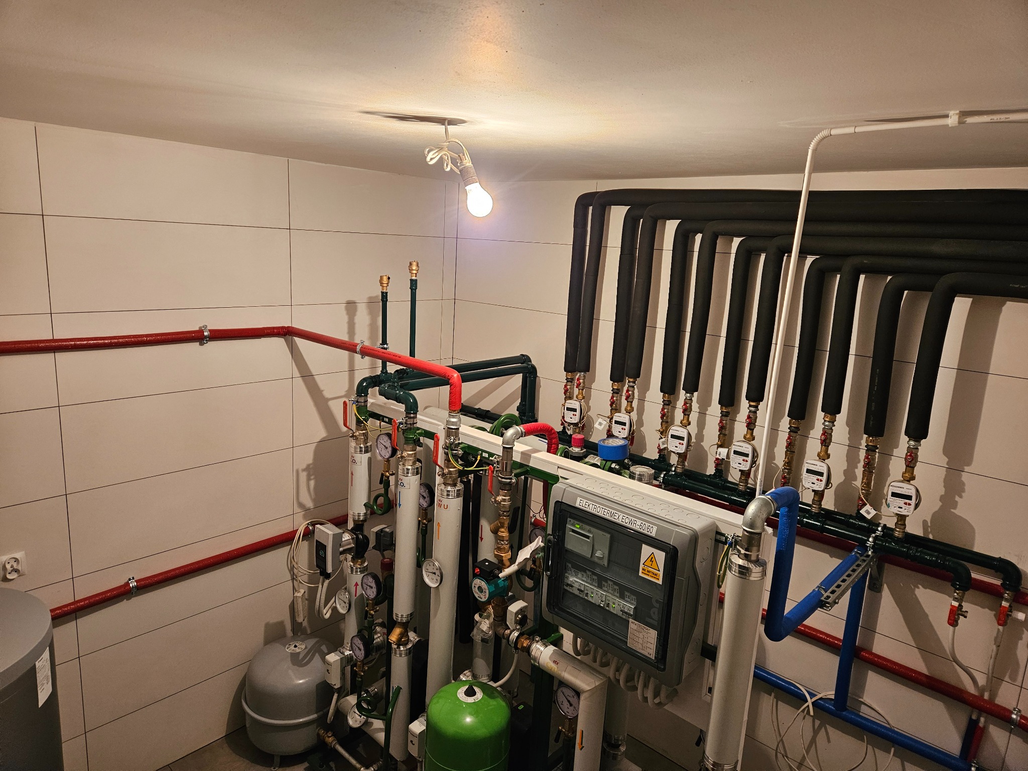Złożona instalacja centralnego ogrzewania z licznymi rurami, zaworami, manometrami i skrzynką elektryczną, oświetlona pojedynczą żarówką na suficie w pomieszczeniu z wyłożonymi kaflami ścianami.