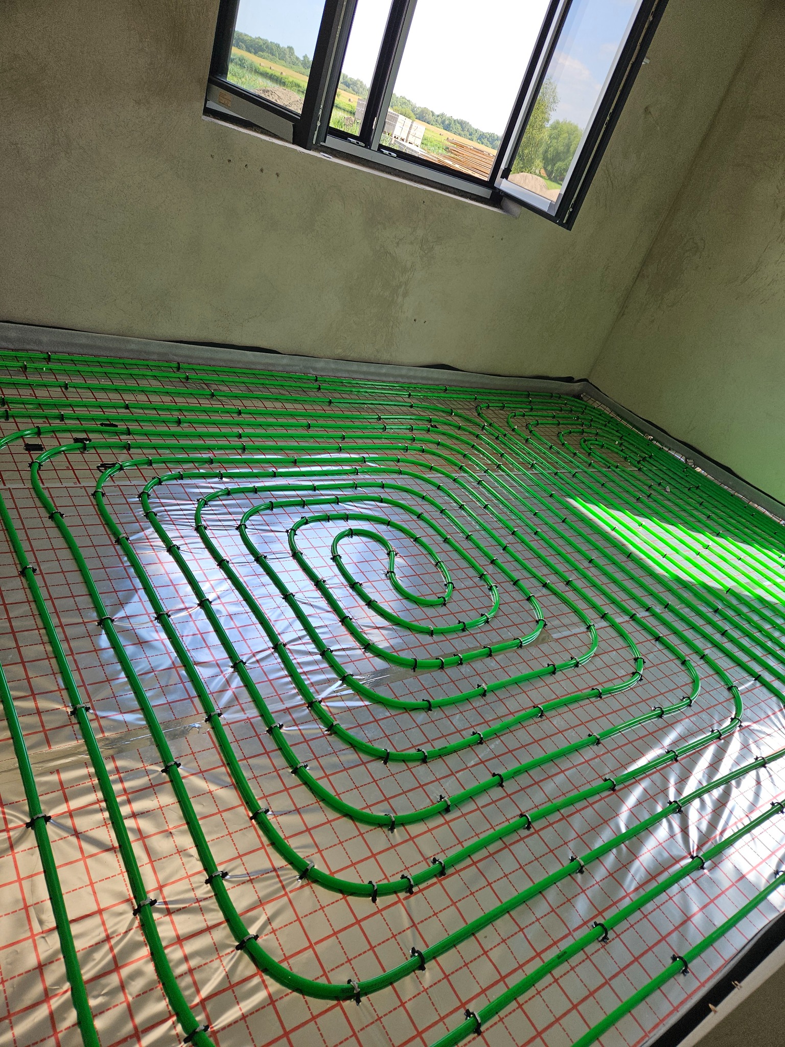 Zielone rury ogrzewania podłogowego ułożone spiralnie na folii z siatką w surowym pomieszczeniu z oknem dachowym.