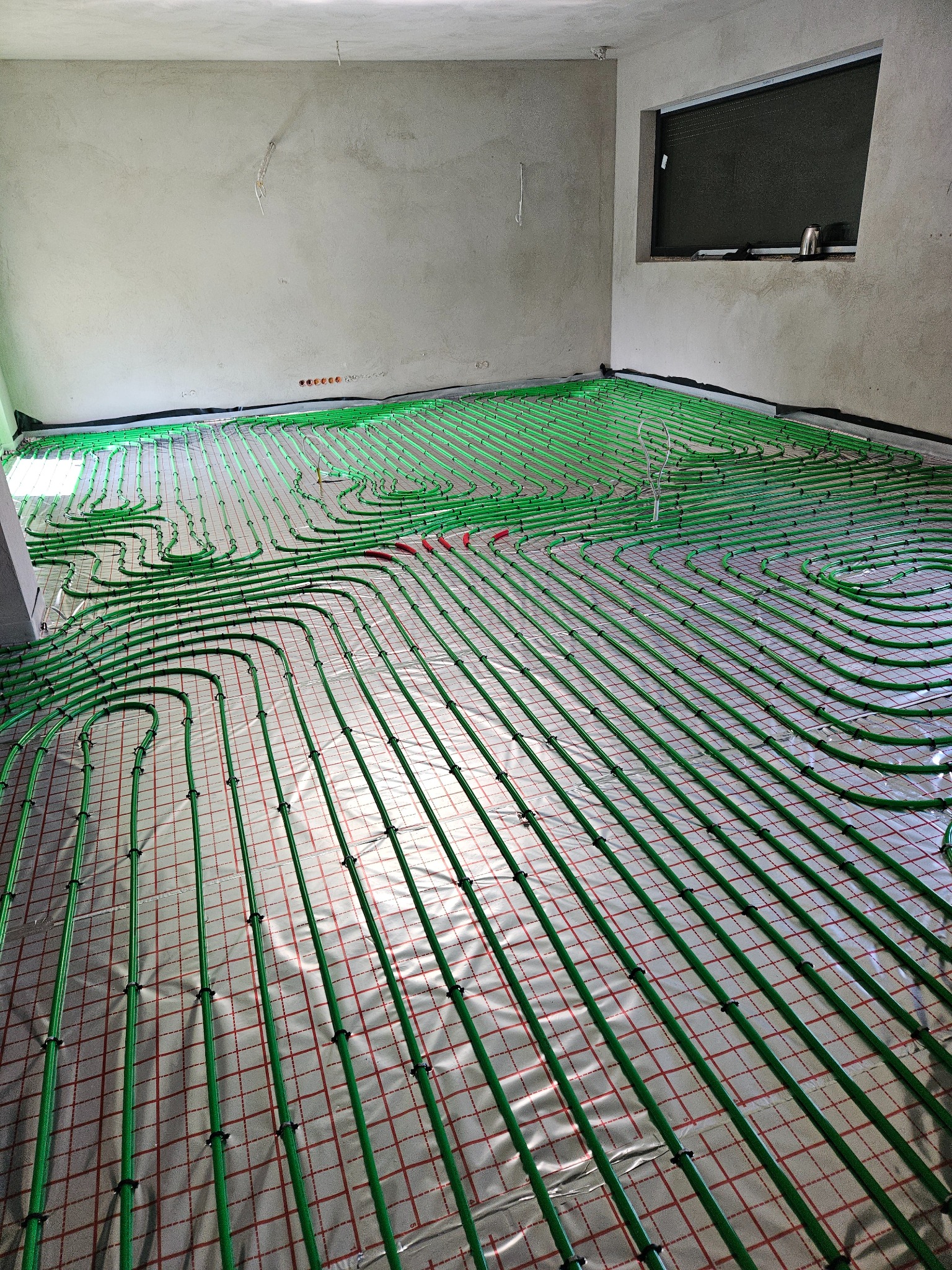 Rozłożona instalacja ogrzewania podłogowego z zielonych rurek na folii z czerwoną kratką w surowym pomieszczeniu przed wylewką.