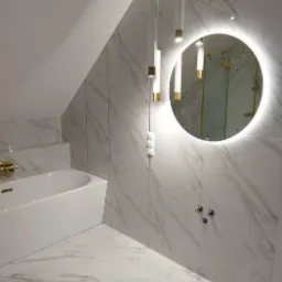 Elegancka łazienka na poddaszu z białymi marmurowymi płytkami, okrągłym lustrem z podświetleniem LED, wiszącą lampą z trzema tubami i białą wanną.