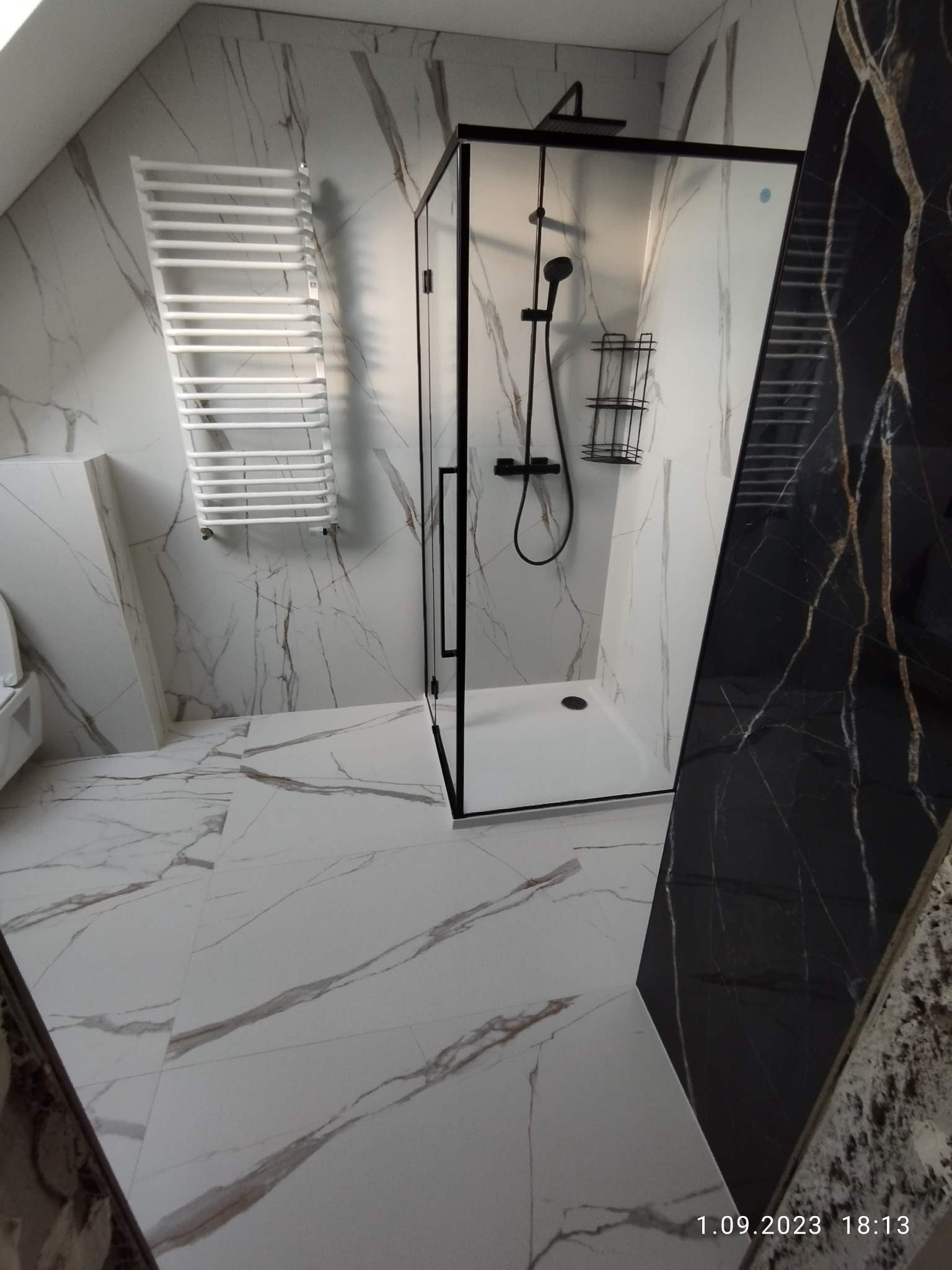 Nowoczesna łazienka z kabiną prysznicową typu walk-in z czarnymi ramami, białymi płytkami imitującymi marmur z szarymi żyłami na ścianach i podłodze, oraz grzejnikiem drabinkowym.