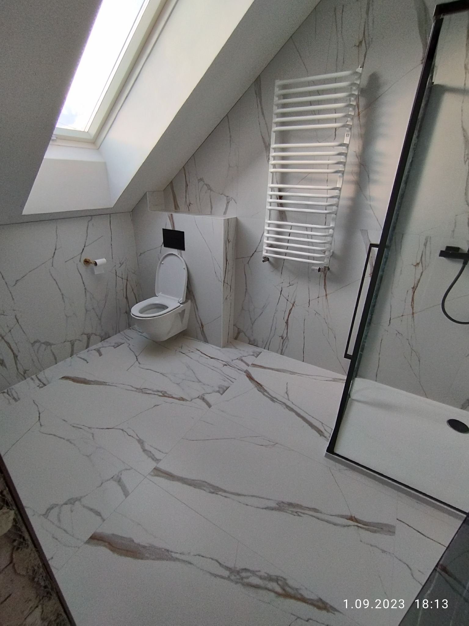 Nowoczesna łazienka na poddaszu z marmurowymi płytkami, toaleta pod skosem dachu, biały grzejnik drabinkowy i kabina prysznicowa z czarnymi ramami.