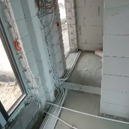 Surowy stan: prowadzenie przewodów w peszlach wzdłuż ścian z betonu komórkowego, widoczne puszki instalacyjne i zwisający pęk kabli.