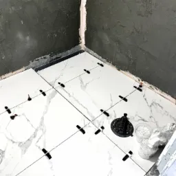 Białe płytki imitujące marmur układane na podłodze w rogu łazienki, z użyciem systemu poziomowania płytek, widoczne przyssawka i pojemniki z klinami.