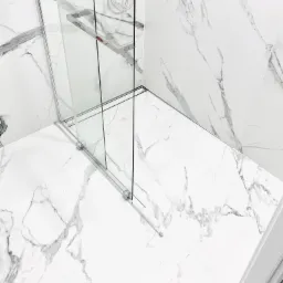 Nowoczesna kabina prysznicowa ze szklanymi drzwiami przesuwnymi i chromowanymi detalami, otoczona białymi płytkami imitującymi marmur z szarymi żyłami, widok z góry na narożnik łazienki.