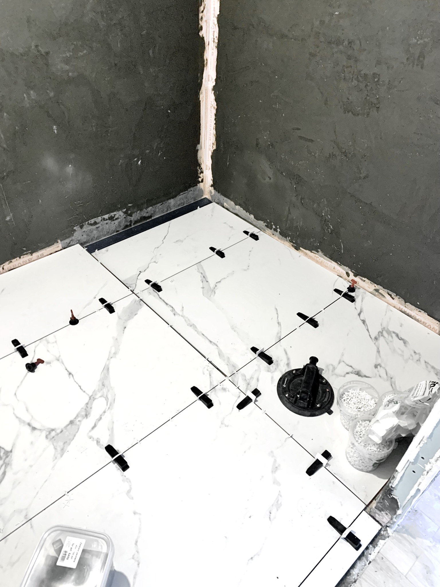 Białe płytki imitujące marmur układane na podłodze w rogu łazienki, z użyciem systemu poziomowania płytek, widoczne przyssawka i pojemniki z klinami.