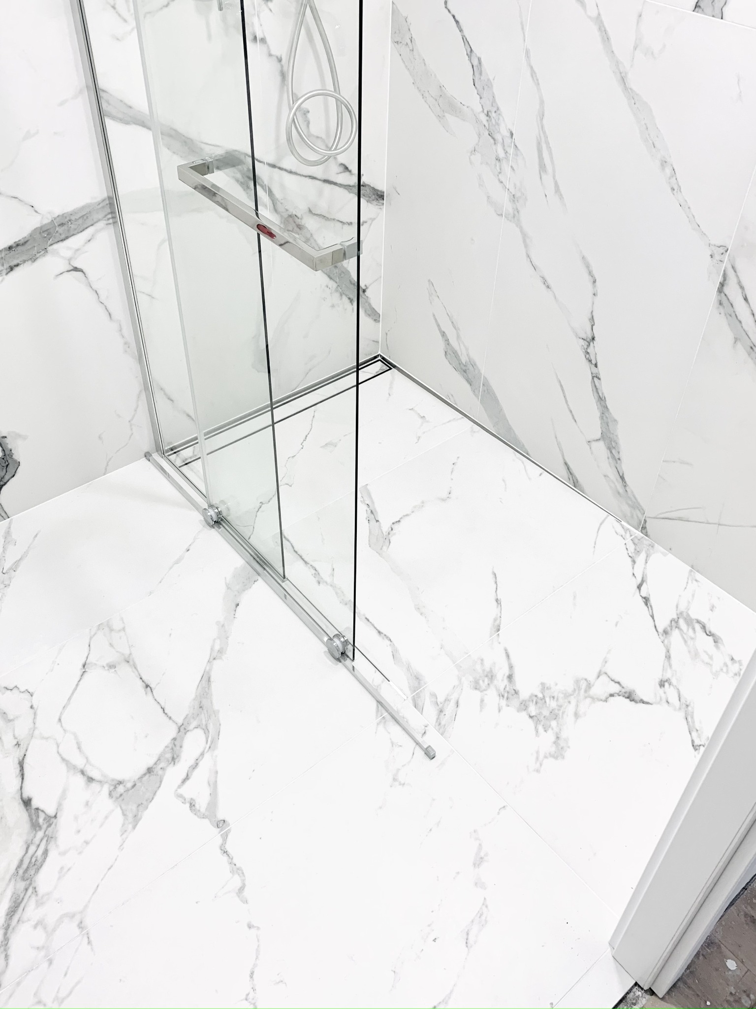 Nowoczesna kabina prysznicowa ze szklanymi drzwiami przesuwnymi i chromowanymi detalami, otoczona białymi płytkami imitującymi marmur z szarymi żyłami, widok z góry na narożnik łazienki.